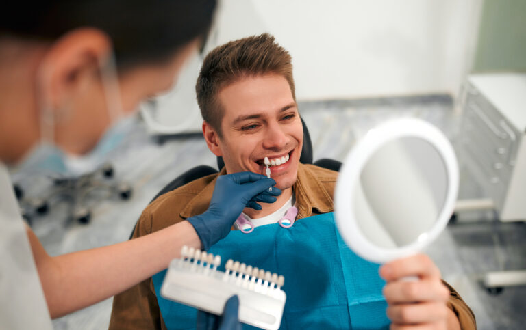 Medicine, dentistry and healthcare concept - closeup of a dentist with tooth color samples choosing a shade for a male patient's teeth in a dental clinic looking at a mirror.