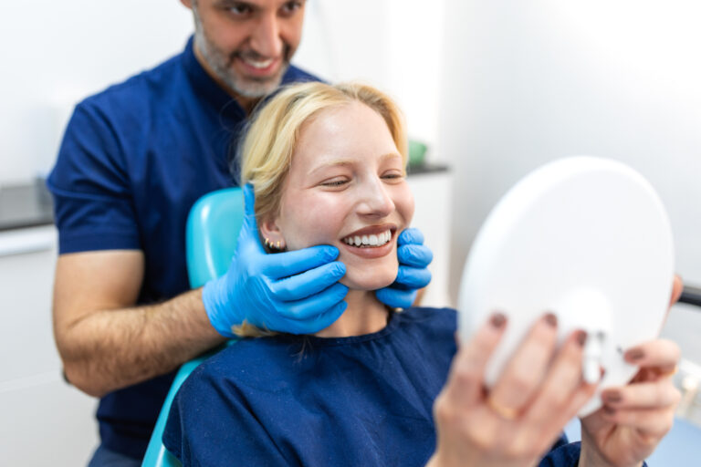 Smiling blonde woman patient happy with her new white teeth and perfect smile in dental mirror at modern dentistry. Professional dentist with gloves behind. Healthcare and medicine concept.