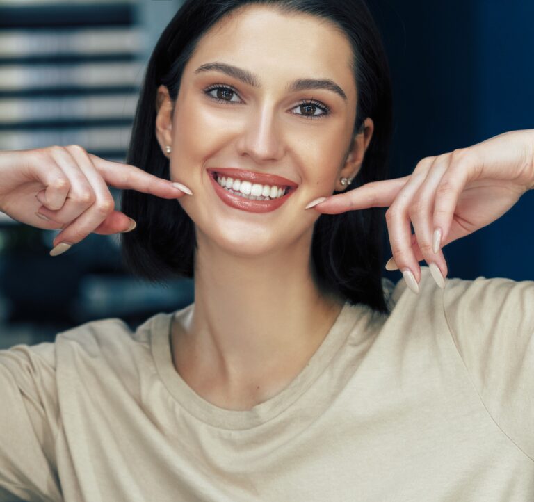 Portrait of a positive attractive girl with natural makeup smiling and showing with index fingers her healthy white teeth. Pretty young woman has joyful expression after makeup.