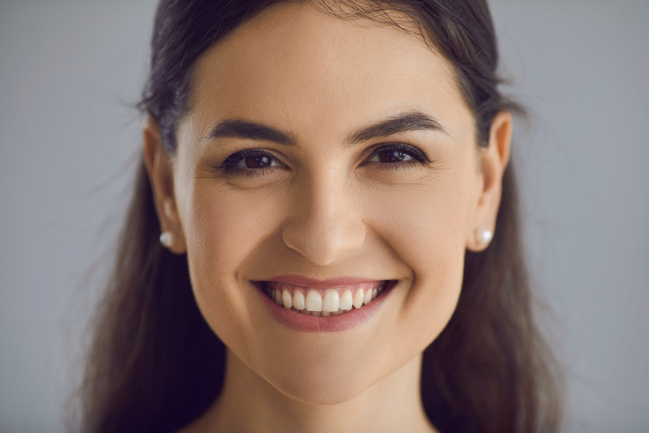 Closeup studio portrait of happy young woman with long dark hair, perfect smile and friendly open face expression looking at camera. Head shot of satisfied patient, customer, client of dental clinic