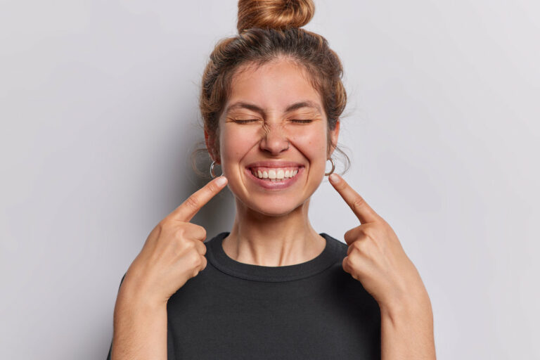Photo of pleasant looking cheerful woman points index fingers at toothy smile shows wwell cared whie even teeth dressed in casual black t shirt isolated over whtie studio background feels happy