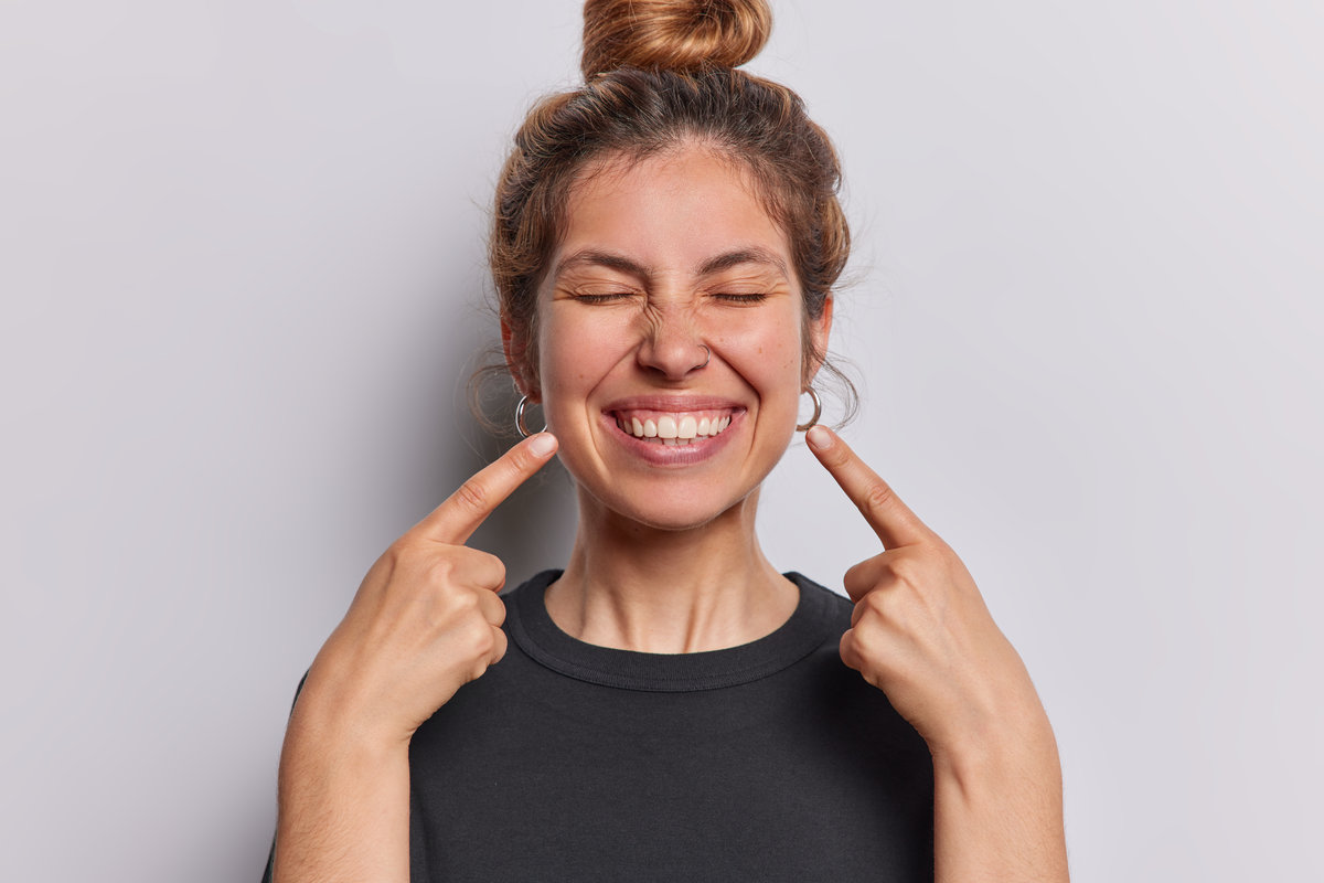 Photo of pleasant looking cheerful woman points index fingers at toothy smile shows wwell cared whie even teeth dressed in casual black t shirt isolated over whtie studio background feels happy