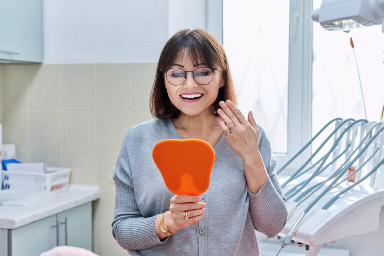 Happy middle aged woman dentist patient with a mirror in her hands looking at her teeth. Treatment, whitening, implants, medicine, health care and beauty smile concept